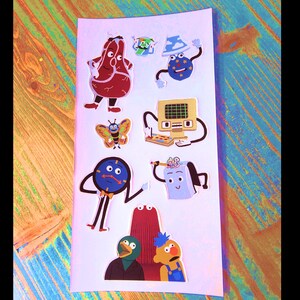 K&ouml;nnte beinhalten: Ein Set mit bunten Comic-Charakter-Stickern mit einer Uhr, einem Computer, einem Schmetterling, einem Notizblock, einem roten Monster, einem blauen Monster und einem Steak.