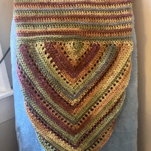 May include: A handmade crochet shawl in a triangular shape, featuring a mix of earthy tones including olive green, brown, and gold. The shawl has a striped pattern with a lacy, openwork design in the lower section. The shawl is draped over a blue surface.