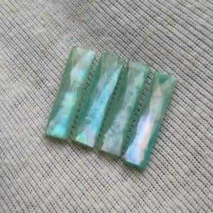 以下が含まれることがあります： 淡い緑色で虹色の真珠のような光沢を持つ、4つの長方形のファセットカットされた宝石。各石は平らで細長い形状をしており、複数の角度のついた表面が光を反射して、きらめく効果を生み出しています。