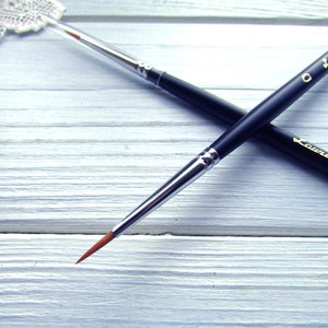 May include: Two black paint brushes with gold lettering, one labeled "Laurel 5/0" and the other "Laurel 0", on a white wooden surface.