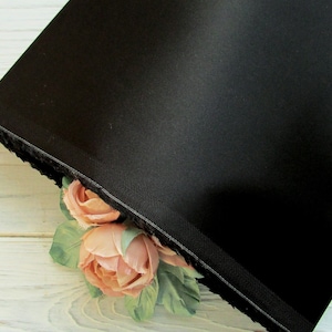 May include: Black fabric with a smooth, satin-like finish. The fabric is folded over and a corner is peeking out from behind two peach-colored fabric roses with green leaves.