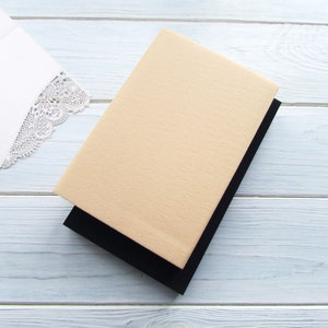 Op de afbeelding: Een beige notitieboek met een zwarte omslag. Het notitieboek is gesloten en ligt op een wit houten oppervlak.