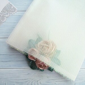 May include: A white sheer fabric with a delicate iridescent sheen and a pink rose flower embellishment. The fabric has a subtle striped pattern and a fringed edge.