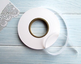 Può includere: Un rotolo di nastro di raso bianco con un'anima di cartone marrone. Un pezzo di nastro è srotolato, appoggiato su una superficie di legno azzurro. Un disegno di carta bianca tagliata al laser è nell'angolo in alto a sinistra.