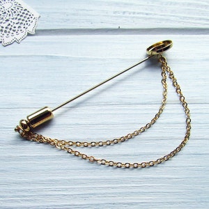 Puede incluir: Un broche de corbata dorado con una cadena. La cadena está unida a una tapa redonda dorada en la parte superior del broche. El broche está sobre una superficie de madera blanca.