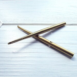 May include: Two gold-colored crochet hooks, one with a curved end and the other with a straight end, are crossed on a white wooden surface.