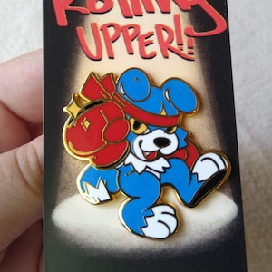 Könnte beinhalten: Anstecknadel mit einem Cartoon-Hund in Blau, Rot und Gold. Der Hund hält einen roten Boxhandschuh und trägt ein rotes Stirnband. Die Anstecknadel trägt den Text "Rolling Upper!" in Rot.