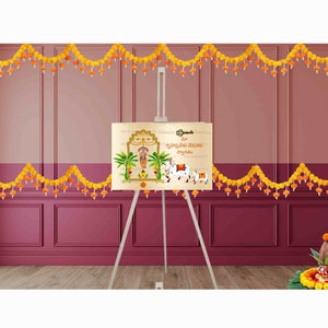 May include: A white easel with a beige poster featuring a colorful illustration of a temple with palm trees and a cow. The poster has text in a foreign language. The easel is in front of a pink wall with yellow flower garlands.