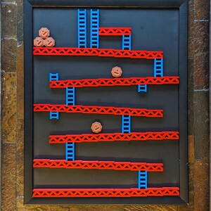 Könnte beinhalten: Ein gerahmtes 3D-Wandbild, das ein klassisches Videospiellevel darstellt. Das Level besteht aus roten und blauen Plastikteilen und zeigt eine braune Figur auf einer Plattform.