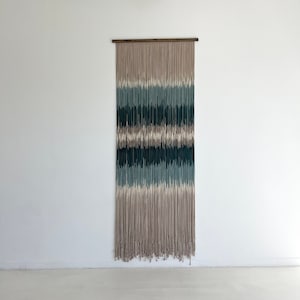 May include: A wall hanging made of yarn in shades of beige, teal, and dark green. The yarn is woven together to create a striped pattern.