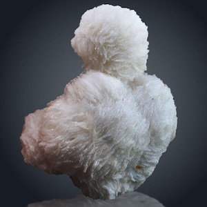 Puede incluir: Un espécimen mineral blanco con una apariencia esponjosa y texturizada. El mineral tiene una forma redondeada con una estructura cristalina en capas. El color es blanco brillante y limpio, y la forma general recuerda a una nube o un racimo de flores. El fondo es un degradado de gris.
