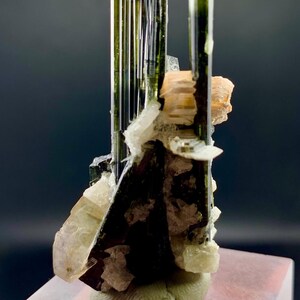 Pode incluir: Um grupo de cristais de turmalina verde e preta sobre uma base branca. Os cristais est&atilde;o dispostos em uma forma&ccedil;&atilde;o vertical, com o cristal maior na parte superior. Os cristais s&atilde;o transl&uacute;cidos e t&ecirc;m um brilho v&iacute;treo.