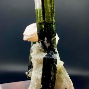 Pode incluir: Um grupo de cristais de turmalina verde e preta com uma matriz de feldspato rosa e branco. Os cristais s&atilde;o afiados e bem formados, e a matriz &eacute; lisa e polida.