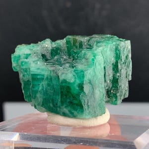 May include: A vibrant, emerald-green crystal cluster. The raw gemstone displays a layered, crystalline structure with a rough, natural texture. The mineral is mounted on a small, white base, showcasing its natural beauty. The background is dark.