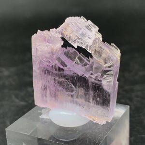 K&ouml;nnte beinhalten: Ein Cluster aus blassrosa, durchscheinenden Kristallen mit einer geschichteten, gestreiften Textur. Die Kristalle sind in einer blockartigen Form angeordnet und werden auf einem klaren Acrylsockel pr&auml;sentiert.