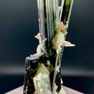 Pode incluir: Um grupo de cristais de turmalina preta com estrias verdes e brancas, colocado sobre uma base branca. Os cristais est&atilde;o dispostos em forma&ccedil;&atilde;o vertical, com o cristal mais alto atingindo aproximadamente 15 cm de altura.