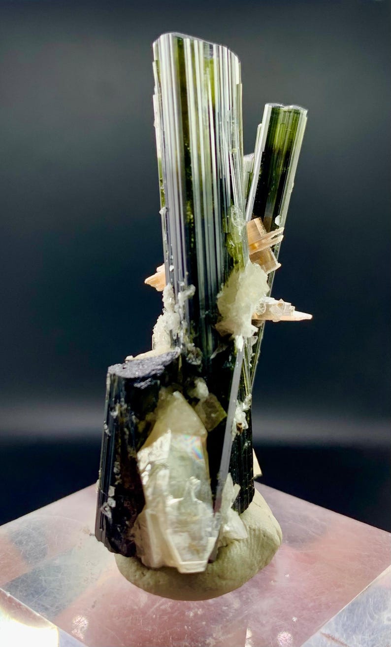 Pode incluir: Um grupo de cristais de turmalina preta com estrias verdes e brancas, colocado sobre uma base branca. Os cristais est&atilde;o dispostos em forma&ccedil;&atilde;o vertical, com o cristal mais alto atingindo aproximadamente 15 cm de altura.