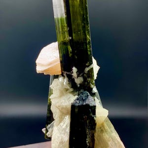 Pode incluir: Um grupo de cristais de turmalina verde e preta com uma matriz de feldspato rosa e branco. Os cristais s&atilde;o afiados e bem formados, e a matriz &eacute; lisa e polida.
