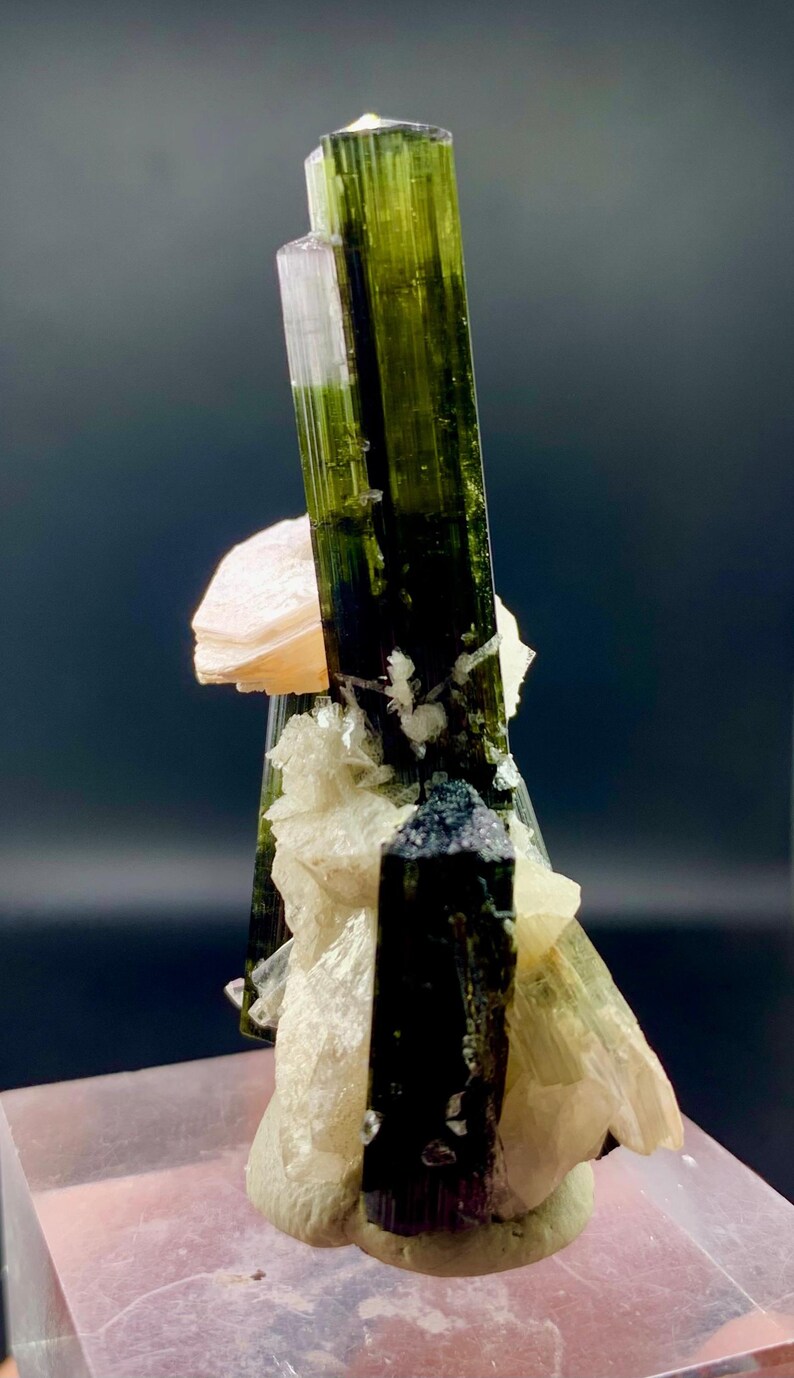 Pode incluir: Um grupo de cristais de turmalina verde e preta com uma matriz de feldspato rosa e branco. Os cristais s&atilde;o afiados e bem formados, e a matriz &eacute; lisa e polida.