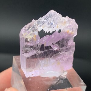K&ouml;nnte beinhalten: Ein Cluster aus durchscheinenden rosa und violetten Kristallen mit einer geschichteten, geometrischen Struktur. Die Kristalle sind auf einem klaren Acrylsockel montiert.