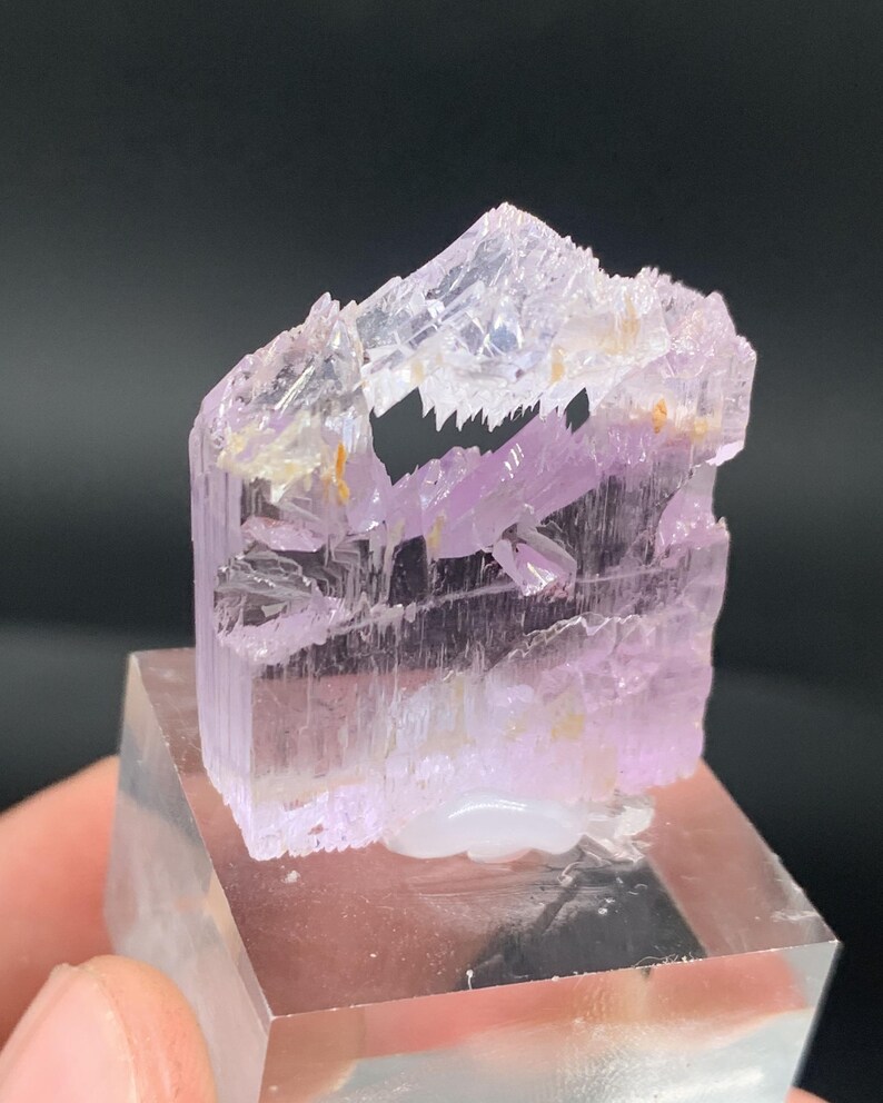 K&ouml;nnte beinhalten: Ein Cluster aus durchscheinenden rosa und violetten Kristallen mit einer geschichteten, geometrischen Struktur. Die Kristalle sind auf einem klaren Acrylsockel montiert.