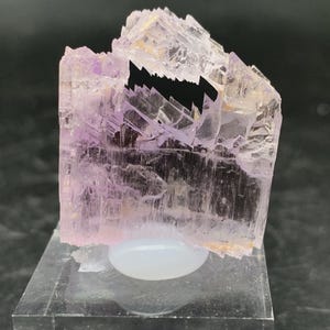 K&ouml;nnte beinhalten: Ein gro&szlig;er, durchscheinender, rosa Kristall mit einer komplexen, facettierten Struktur. Der Kristall wird auf einem klaren Acrylsockel pr&auml;sentiert.