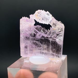 K&ouml;nnte beinhalten: Ein durchscheinender rosa Kristall mit einer einzigartigen, unregelm&auml;&szlig;igen Form. Der Kristall wird auf einem klaren Acrylsockel pr&auml;sentiert.