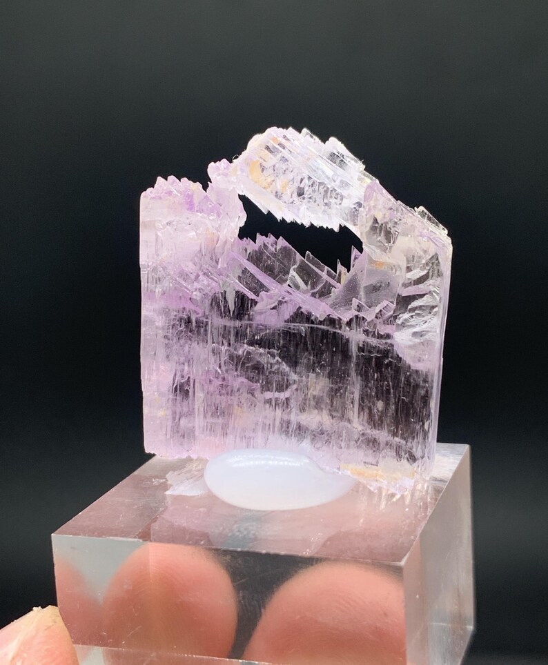 K&ouml;nnte beinhalten: Ein durchscheinender rosa Kristall mit einer einzigartigen, unregelm&auml;&szlig;igen Form. Der Kristall wird auf einem klaren Acrylsockel pr&auml;sentiert.