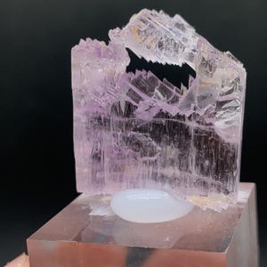 K&ouml;nnte beinhalten: Ein durchscheinender rosa Kristall mit einer einzigartigen, geschichteten Struktur. Der Kristall wird auf einem klaren Acrylsockel pr&auml;sentiert.