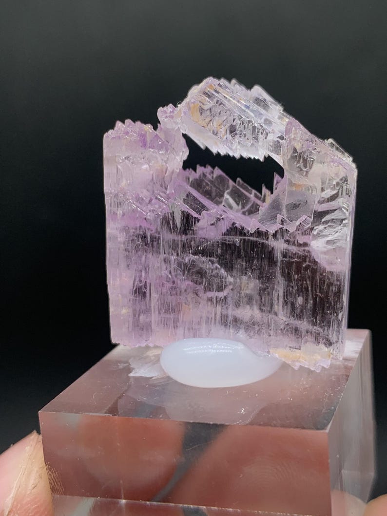 K&ouml;nnte beinhalten: Ein durchscheinender rosa Kristall mit einer einzigartigen, geschichteten Struktur. Der Kristall wird auf einem klaren Acrylsockel pr&auml;sentiert.