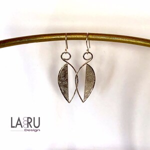 Peut inclure: Boucles d'oreilles argentées en forme de feuille avec un design bicolore. Un côté est gris métallique texturé, l'autre est blanc. Les boucles d'oreilles sont suspendues à des crochets argentés et sont présentées sur une barre incurvée dorée. La marque "LA RU Design" est visible.