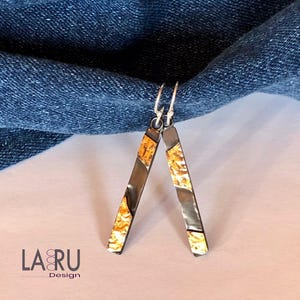 May include: A pair of silver earrings with a rectangular design. The earrings have a dark grey metal frame with gold-coloured accents. The earrings are hanging against a blue denim background. The earrings are approximately 5 cm long.