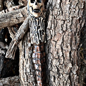 May include: A carved wooden skull-shaped object with a detailed bone design. The skull is beige with dark eye sockets and an open mouth. The object has a dark brown, textured base and is set against a backdrop of tree bark and branches.