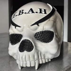 May include: A white skull-shaped helmet with black detailing and the letters "E.B.A.H" across the top. The eye sockets are covered with a black mesh, and the jaw features prominent fangs. The helmet is likely for decorative or costume purposes.