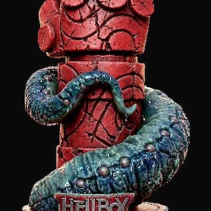 May include: A red Hellboy statue featuring a large hand with two fingers raised, a cylindrical body, and a blue tentacle. The base has the Hellboy logo and a decorative design. The statue is set against a black background.