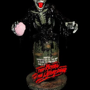 May include: A detailed figurine of a zombie emerging from a green barrel. The zombie has a decaying appearance with exposed bones, a red mouth, and a pink brain. The base features the text "The Return of the Living Dead."