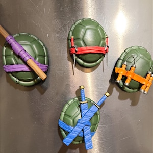May include: Four Teenage Mutant Ninja Turtles themed magnets. Each magnet features a green turtle shell design with different colored accessories: purple, red, orange, and blue. The accessories include weapons and headbands.