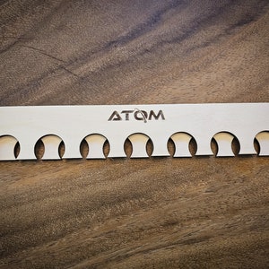 May include: A wooden organizer with the word "ATOM" engraved on it. The organizer has ten round cutouts for holding small items.