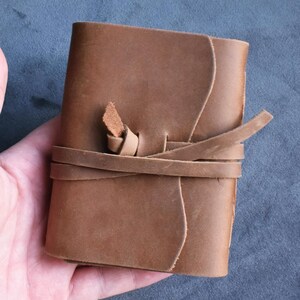 Może przedstawiać: Mały, brązowy skórzany notatnik z zapięciem na sznurek. Notatnik jest trzymany w dłoni osoby na ciemnoszarym tle. Skóra ma naturalny, teksturowany wygląd.