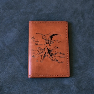 Könnte beinhalten: Ein brauner Leder-Reisepasshalter mit einer gravierten Abbildung eines Berges und eines Drachen, der darüber fliegt. Der Text "The Lonely Mountain" ist auf der Vorderseite eingraviert.