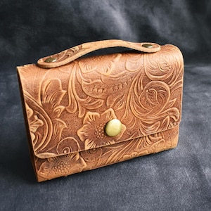 May include: A small, brown leather purse with a floral embossed design. The purse has a handle and a brass-colored clasp. The overall design is a classic, vintage style. The purse is sitting on a dark gray surface.