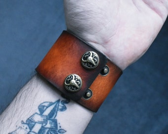 May include: A brown leather cuff bracelet with two decorative metal buttons. The buttons have a black and gold design.