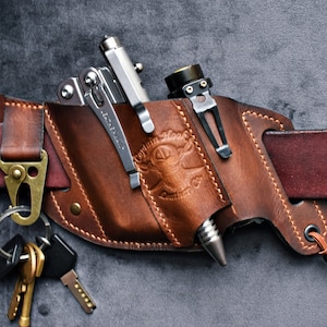 May include: A brown leather belt holster with a multi-tool, flashlight, and pen. The holster is attached to a brown leather belt and has a key ring with keys attached.