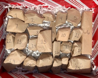 May include: A rectangular box with red and white striped edges containing twenty-four individual items wrapped in brown paper, each marked with a handwritten number from one to twenty-four, nestled in white shredded paper filler.