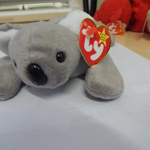 May include: Gray plush koala bear with black eyes and a black nose. The koala is lying on its belly with its paws outstretched. The bear has a red Ty tag attached to its back that says "Beanie Babies".