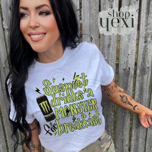 May include: White t-shirt with the text "Suspect drinks a MONSTER for breakfast" in green and black. The shirt features a graphic of a Monster energy drink can. The shirt is worn by a person.
