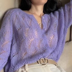 May include: A light purple, open-weave cardigan with long sleeves. The cardigan is buttoned at the chest and worn with cream-colored pants and a light-colored belt. The cardigan has a delicate, lacy pattern.