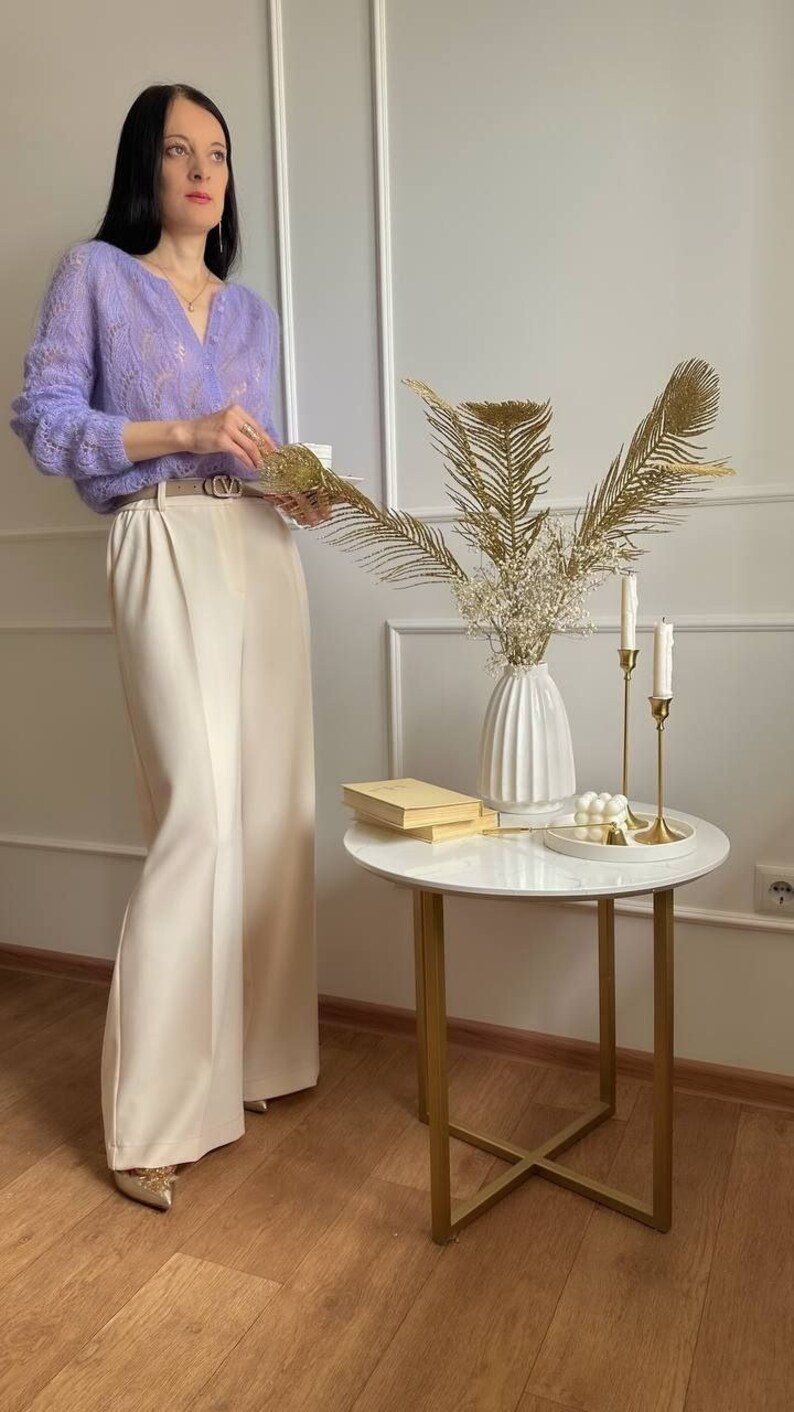 May include: A woman wearing a light purple, open-knit sweater and cream-colored wide-leg pants. A small, round side table with gold legs holds a white vase with dried flowers, candles, and books. The overall aesthetic is minimalist and elegant.