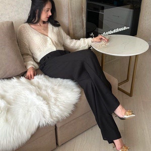 May include: A woman wearing a cream-colored, open-knit cardigan and black trousers. She is seated on a beige sofa with a white faux fur throw. Gold-colored heels with floral embellishments complete the outfit. A small, round table is nearby.
