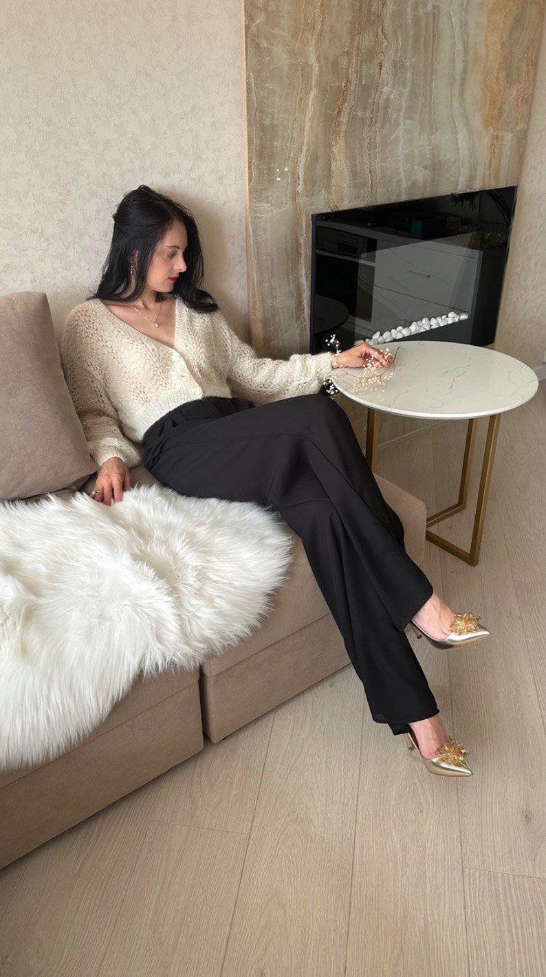 May include: A woman wearing a cream-colored, open-knit cardigan and black trousers. She is seated on a beige sofa with a white faux fur throw. Gold-colored heels with floral embellishments complete the outfit. A small, round table is nearby.
